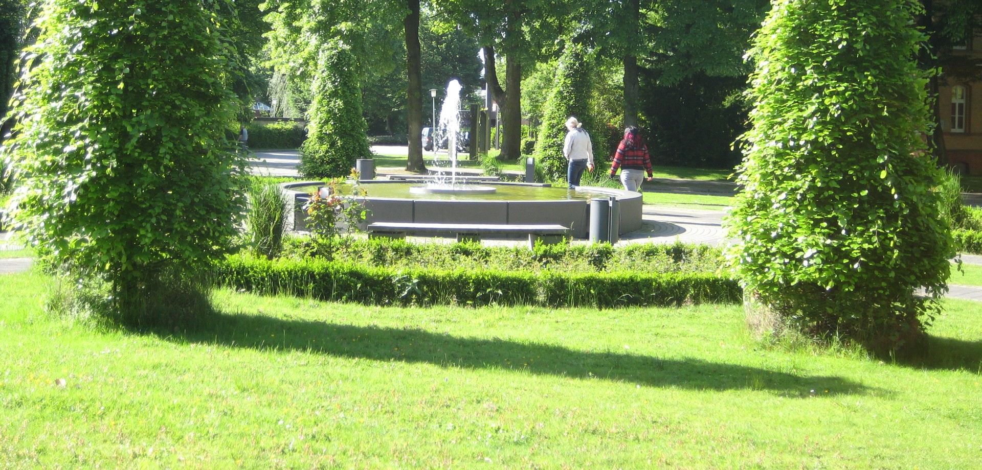 Zwei Frauen gehen an einem großen, sprudelnden Brunnen vorbei  Sie sind von hinten fotografiert. Der Brunnen ist von grünen Bäumen umgeben.