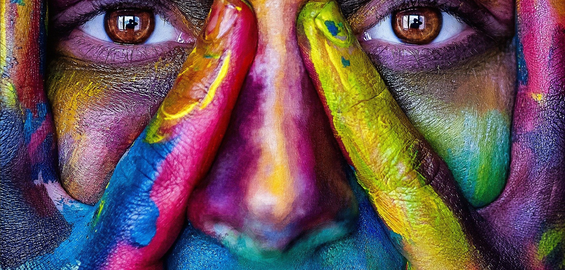 In Großaufnahme das Gesicht einer Frau, deren Gesicht und Hände kunterbunt in leuchtenden Farben angemalt sind