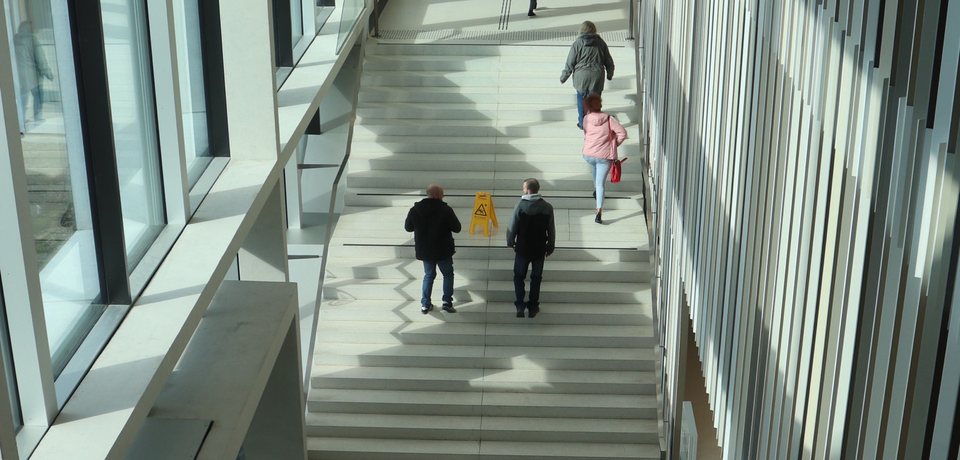 Auf deiner breiten Treppen, in einem lichten, von der Sonne beschienenen Treppenflur, gehen mehrere jüngere Menschen die Stufen hinauf.