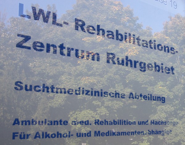 Auf eine Glastür ist ein gedruckter Hinweis auf das LWL-Rehabilitationszentrum zu lesen. In der Tür spiegeln sich grüne Bäume